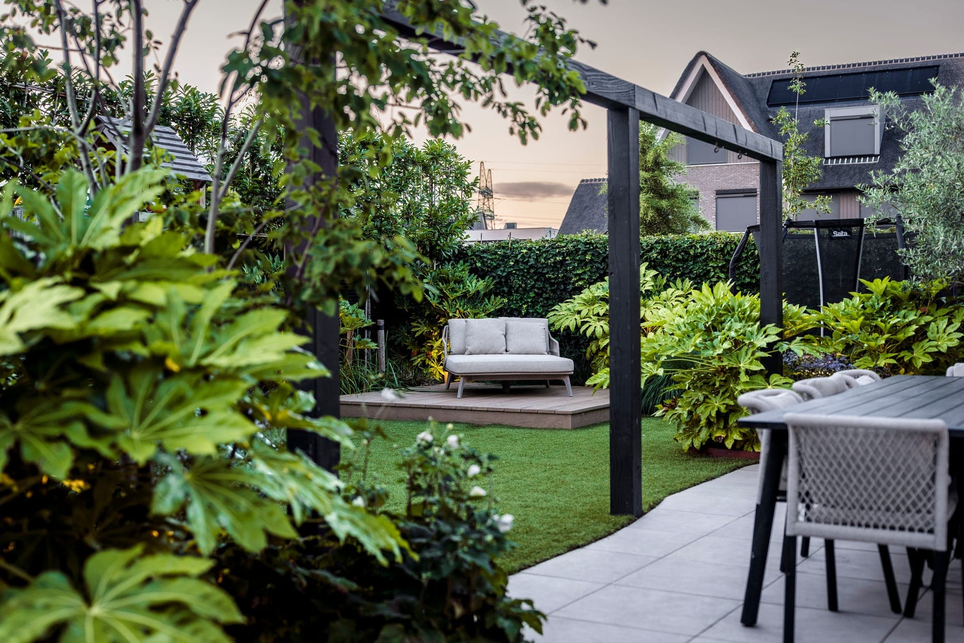 Tuinoverzicht in de schemer met pergola-poort, daybed en kunstgras