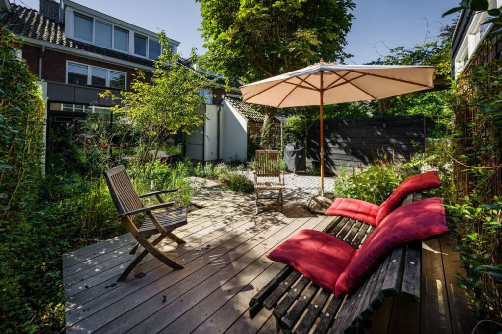 Een houten terras met een rode parasol en kussens. In de omgeving zijn planten, een schutting en een achtergevel van een huis