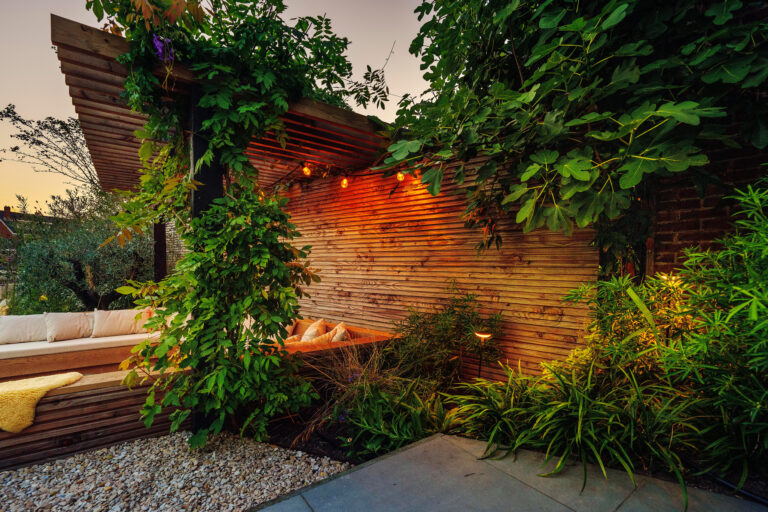 Een houten terras met een comfortabele bank en kussens, omringd door weelderige planten. Verlichte achterwand.